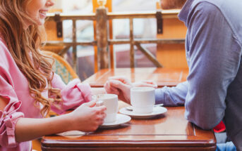 A woman and man are sitting at a table in a cafe, each holding a cup of coffee. The woman is wearing a pink blouse and smiling, and the man is in a blue shirt. They are engaged in conversation, and the background shows an outdoor seating area with blurred details.