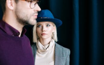 A woman with light hair, wearing a blue hat and a grey checkered blazer over a beige turtleneck, gazes forward with a serious expression. A man wearing glasses and a purple shirt is in the foreground, slightly out of focus, facing away from her.