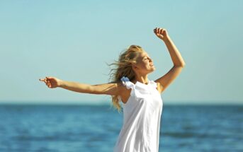 A person with long blonde hair is wearing a white dress and standing with their arms raised, enjoying the sunshine by the ocean. The sky is clear blue, and the sea is calm in the background.