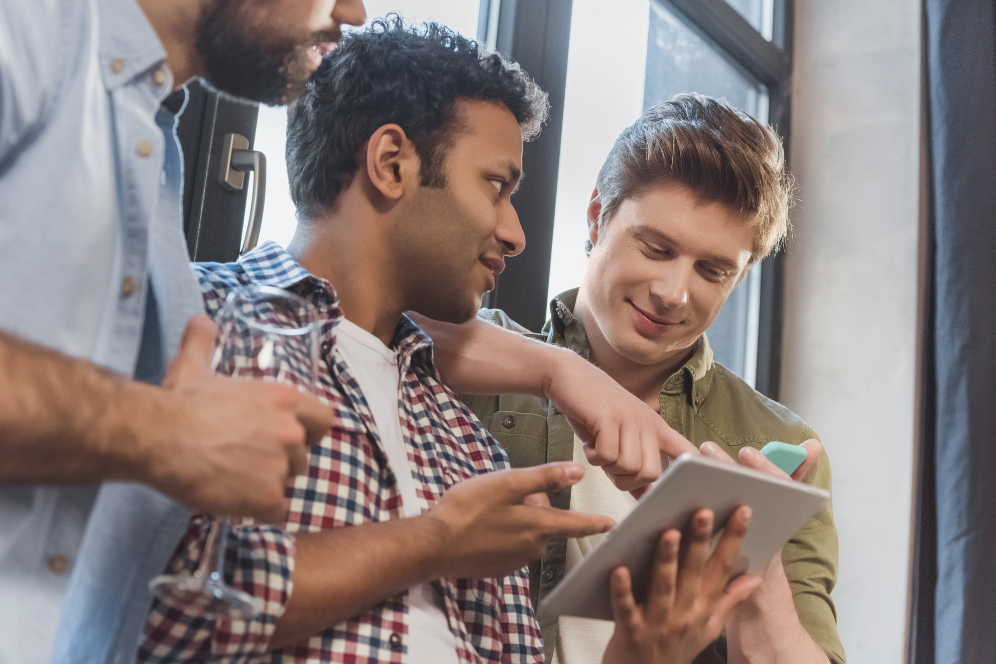 Three men are gathered around a tablet, engaged in discussion. One man, holding a glass of water, is talking; another is tapping on the tablet screen, and the third is smiling while holding a smartphone. They are indoors, near a large window.