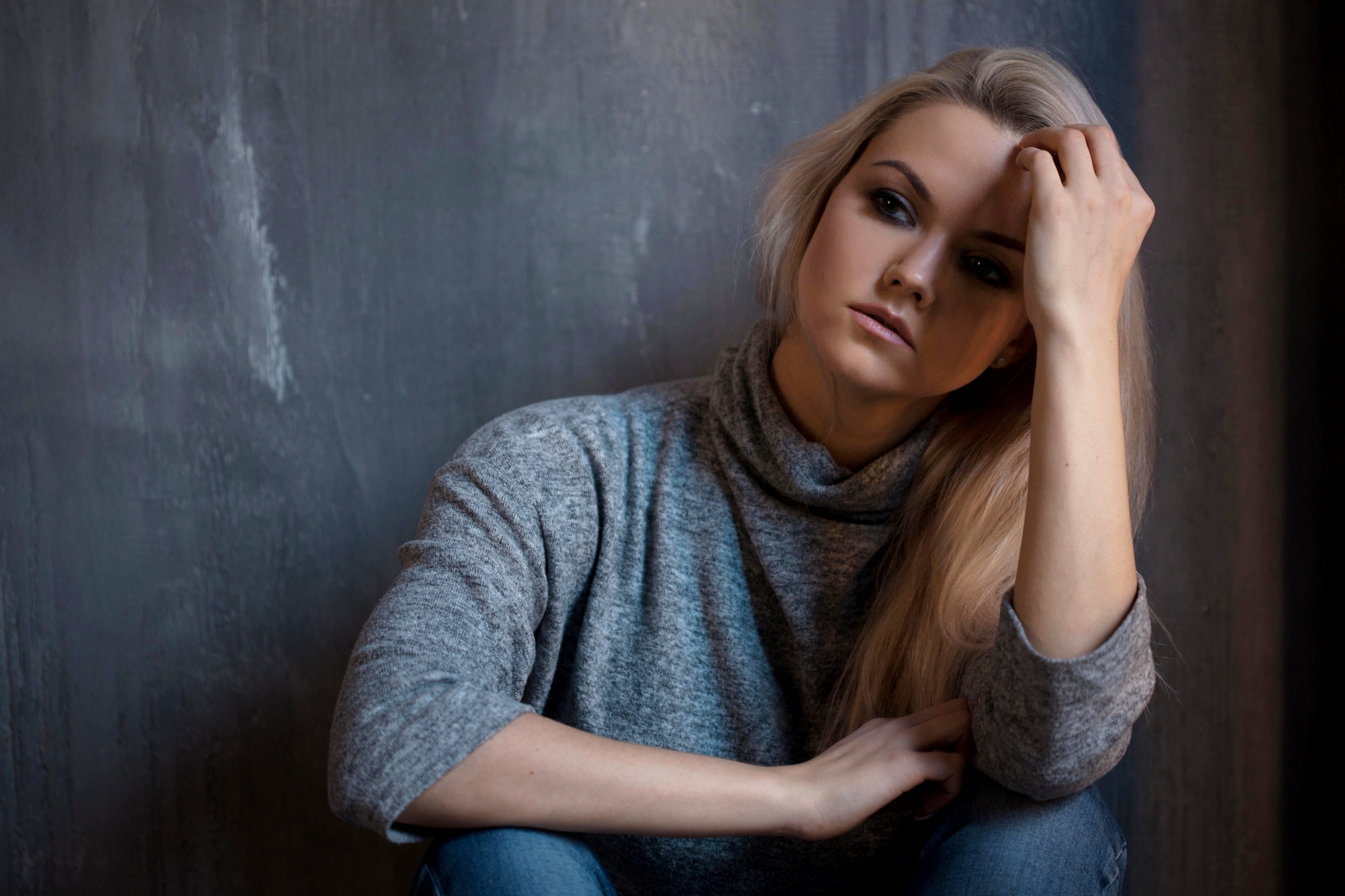 A woman with long blonde hair is sitting against a textured gray wall. She wears a light gray turtleneck sweater and jeans, gazing thoughtfully with one hand resting on her head and the other on her knee. The lighting is soft and subdued.