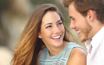 A woman with long brown hair and a man with short brown hair share a joyful moment, smiling at each other. The woman is wearing a sleeveless blue top, and the man is wearing a white shirt. The background is blurred, suggesting an outdoor setting.