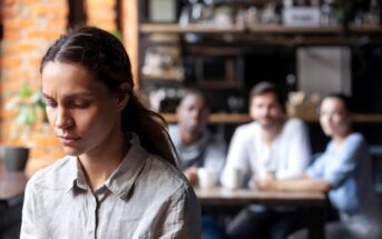 A woman with a sad expression sits alone in the foreground of a café, looking down. In the background, three people sit together at a table, with two of them looking in her direction. The scene suggests a sense of isolation and contemplation.