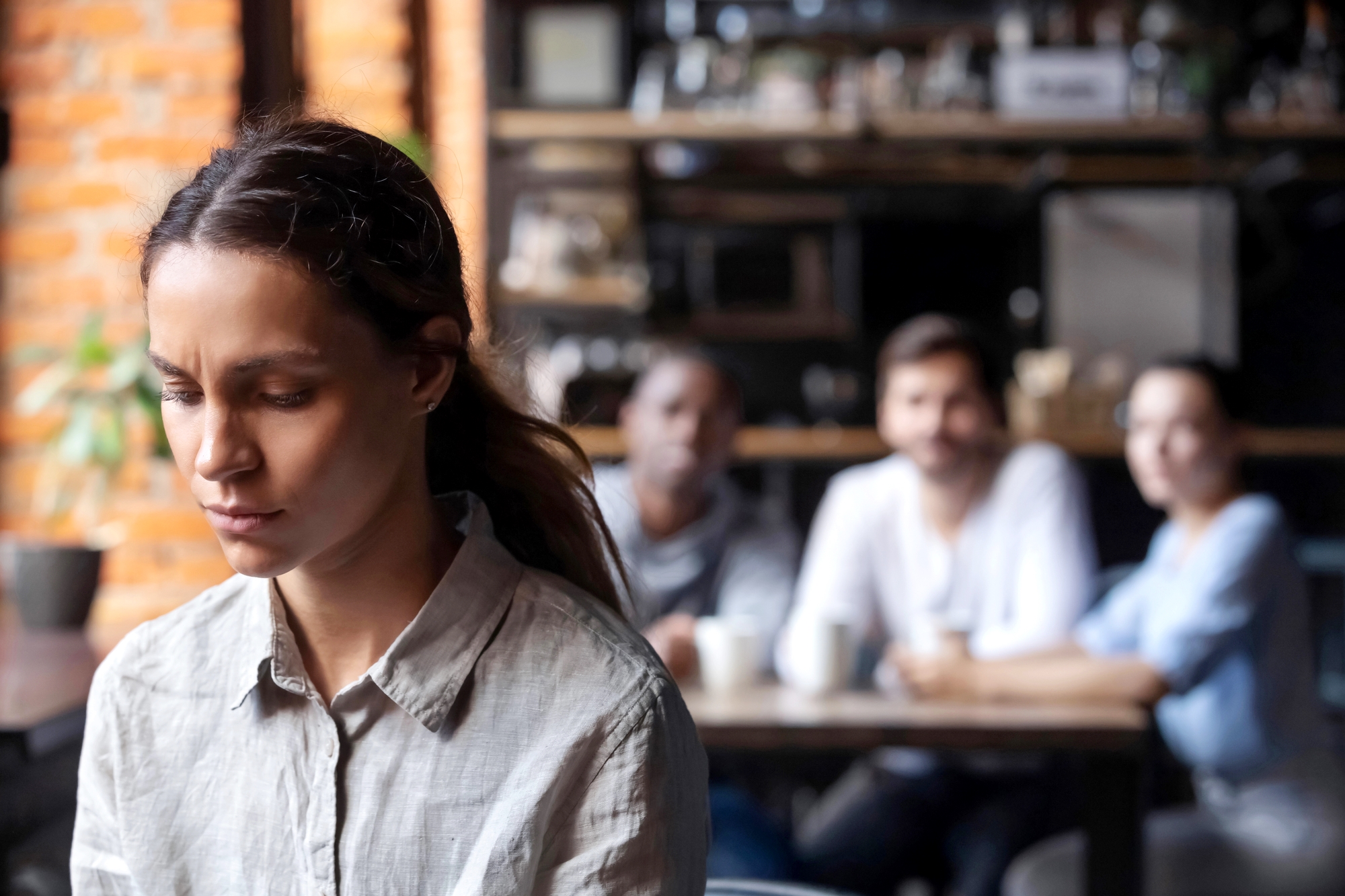 A woman with a sad expression sits alone in the foreground of a café, looking down. In the background, three people sit together at a table, with two of them looking in her direction. The scene suggests a sense of isolation and contemplation.