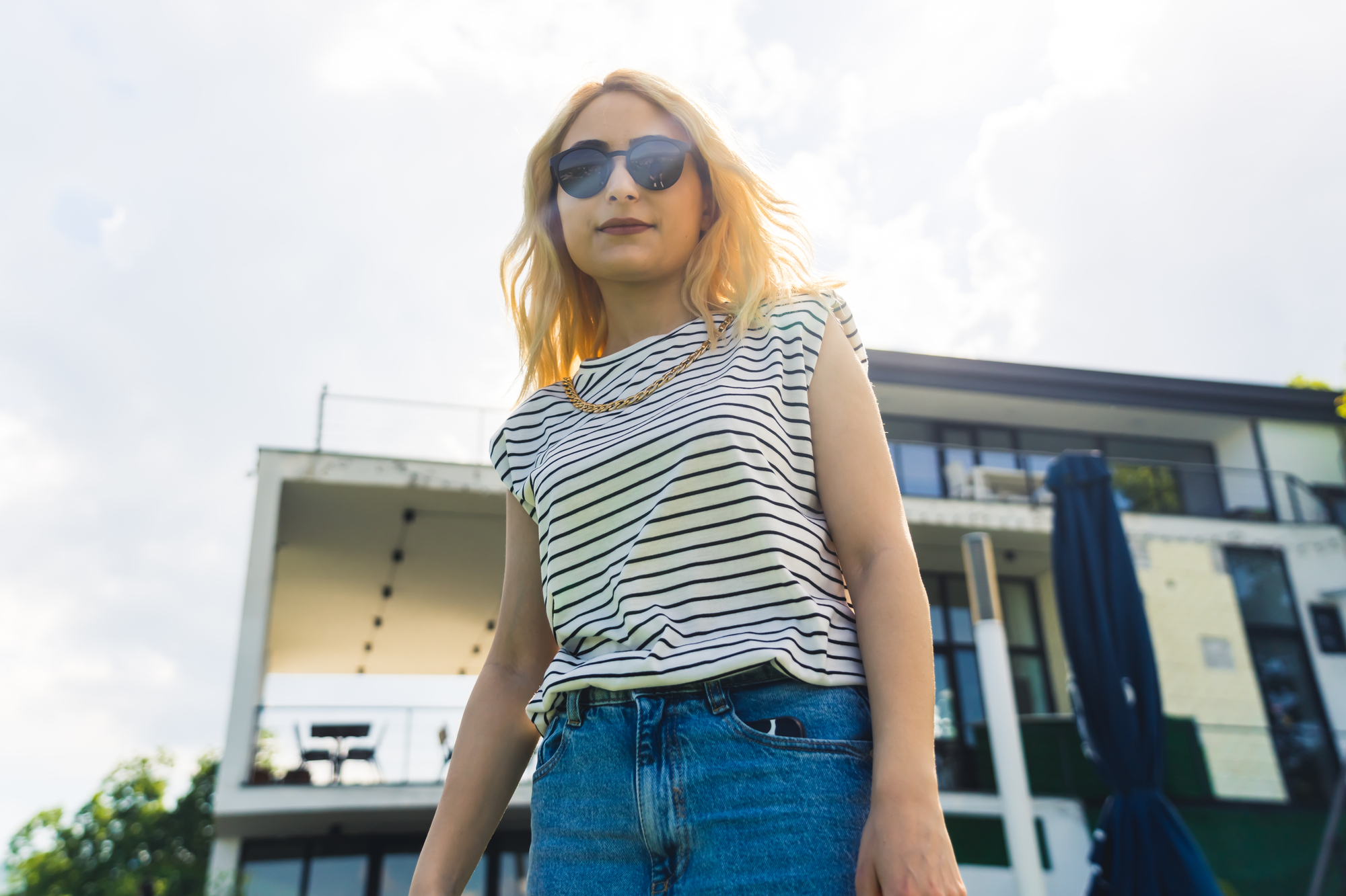 A woman with long blonde hair wearing sunglasses and a striped shirt stands confidently outdoors. She has one hand in the pocket of her blue jeans. In the background, there is a modern building with large windows and a patio area. The sky is partly cloudy.
