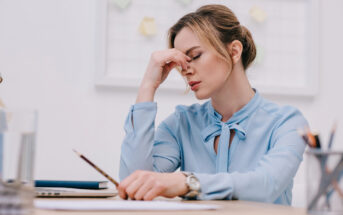 A woman wearing a light blue blouse sits at a desk with her eyes closed, pinching the bridge of her nose in frustration or fatigue. There are papers, a pencil, a laptop, and office supplies on the desk. The background shows a whiteboard with sticky notes.