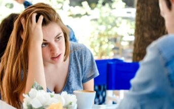 A woman with brown hair rests her head on her hand, looking intently at another person, who is not fully visible, during a conversation at an outdoor café. A coffee cup and a small vase of flowers are on the table in front of her.