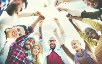 A diverse group of people stands in a circle with their hands raised and fists clenched, smiling joyfully. They appear cheerful and united, captured from a low-angle view against a clear sky backdrop. They are casually dressed, showcasing various styles.