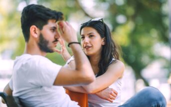 A woman and a man are sitting outdoors on a bench, engaged in a conversation. The woman is leaning forward with her arms resting on her knees, wearing a white sleeveless top and sunglasses on her head. The man in a white t-shirt is touching his forehead. Trees are in the background.