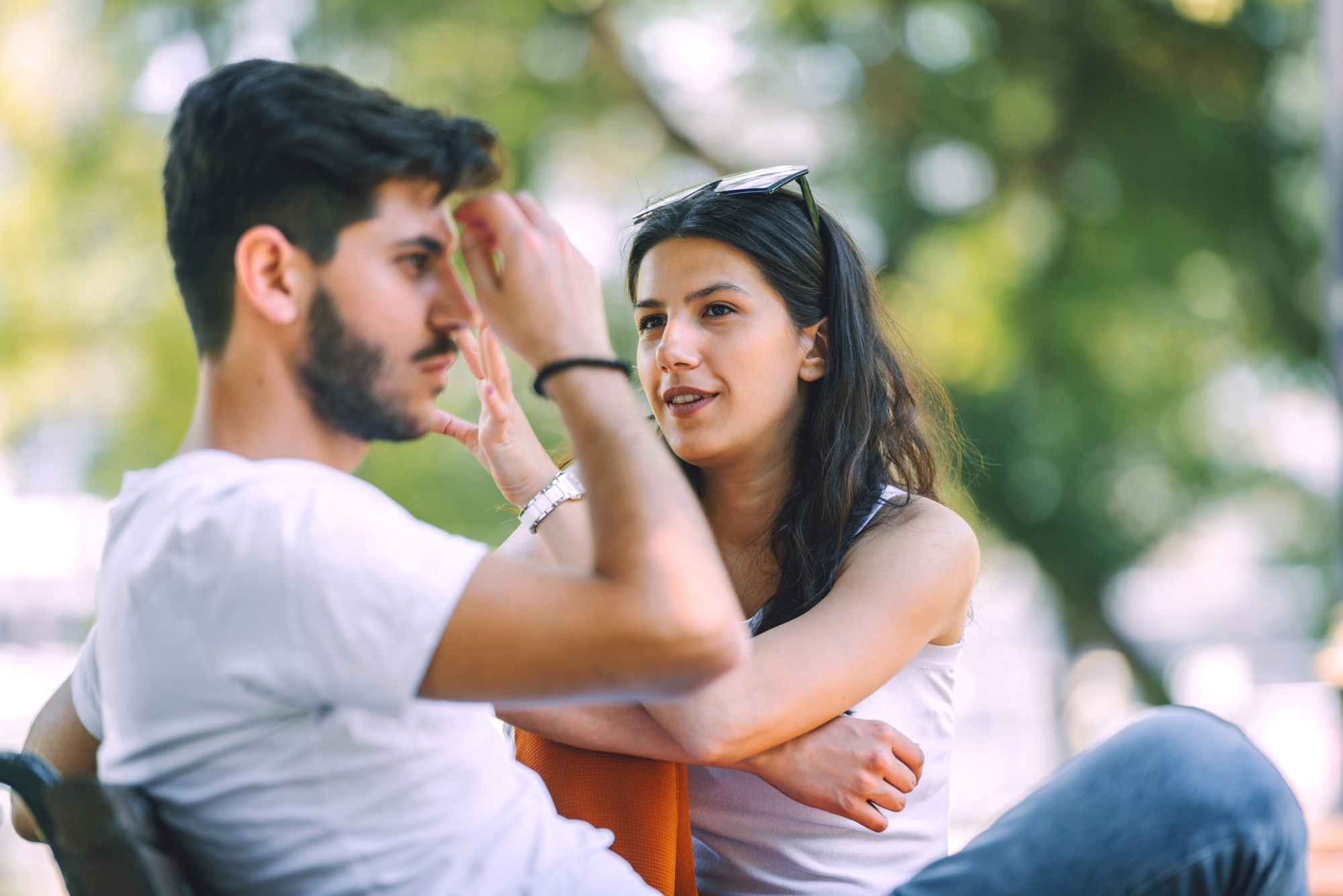 A woman and a man are sitting outdoors on a bench, engaged in a conversation. The woman is leaning forward with her arms resting on her knees, wearing a white sleeveless top and sunglasses on her head. The man in a white t-shirt is touching his forehead. Trees are in the background.