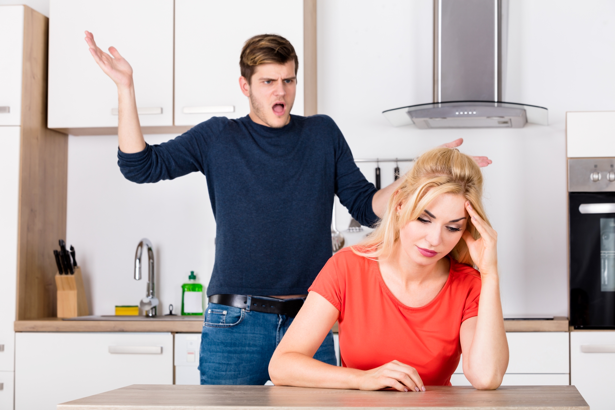 A man with a frustrated expression is standing in a kitchen, gesturing with his hands. A woman with a downcast expression sits at a table in the foreground, leaning her head on one hand. The scene suggests they are having a disagreement.