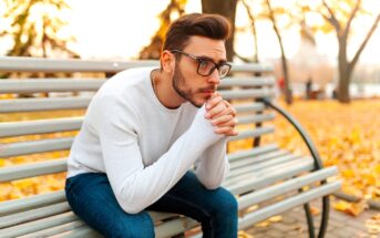 A man with glasses, wearing a white sweater and blue jeans, sits on a park bench in an autumn setting. He is resting his chin on his clasped hands, looking thoughtful. Fallen leaves surround the bench and trees with yellow foliage are visible in the background.