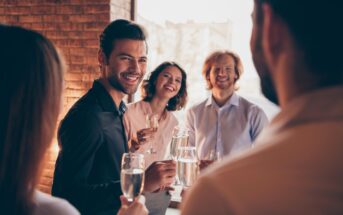 A group of four people, three men and one woman, are standing in a warmly lit room and smiling while holding glasses of champagne. The background includes a brick wall and large windows showing an outdoor view. They appear to be engaged in a cheerful conversation.