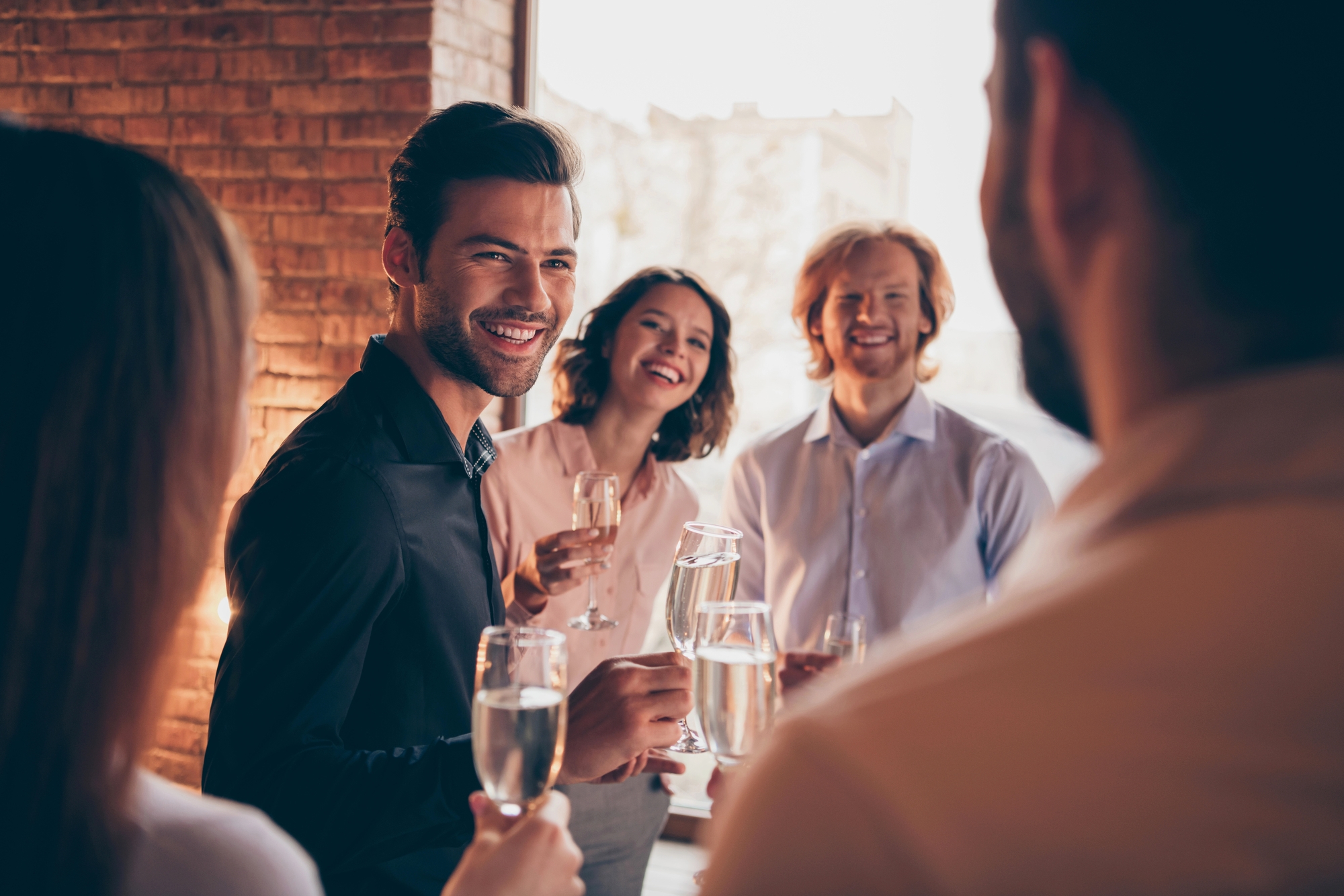 A group of four people, three men and one woman, are standing in a warmly lit room and smiling while holding glasses of champagne. The background includes a brick wall and large windows showing an outdoor view. They appear to be engaged in a cheerful conversation.