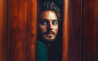 A man with wavy hair and a beard looks through a narrow gap between wooden panels, his face softly lit and eyes focused, creating a contemplative and slightly mysterious mood.