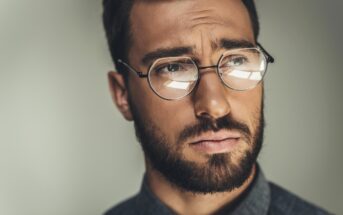 A close-up of a man with brown hair and a beard wearing round glasses and a serious expression. He has a contemplative look in his eyes and is dressed in a dark shirt, with a blurred neutral background.