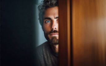 A man with blue eyes and a beard looks through a narrow gap between a wooden door and a wall, with only part of his face visible in soft light.
