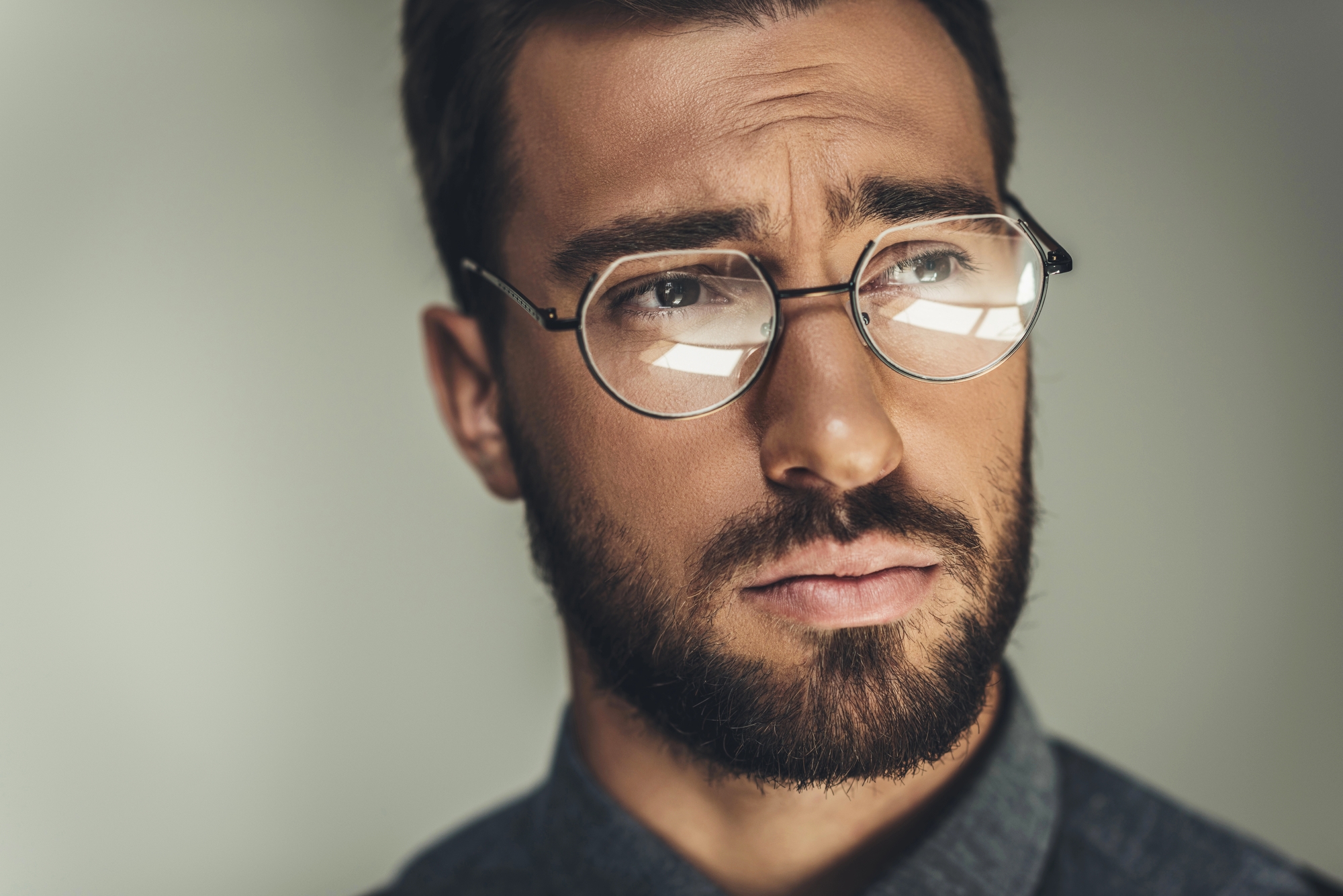 A close-up of a man with brown hair and a beard wearing round glasses and a serious expression. He has a contemplative look in his eyes and is dressed in a dark shirt, with a blurred neutral background.