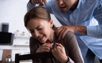 A woman sits in a chair, looking distressed and upset, while a man stands behind her, gripping her shoulders and shouting aggressively. Both are indoors in a kitchen setting, with blurry shelves and items visible in the background.