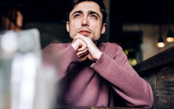 A man with short dark hair is sitting at a wooden table indoors, resting his chin on his clasped hands and looking thoughtfully into the distance. He is wearing a pink knit sweater, and the background is softly blurred with warm lighting.