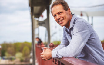 A smiling man in a light gray sweater leans on the railing of a bridge. The bridge structure is visible behind him, and there is a scenic view with greenery and water in the background. The sky is slightly overcast.