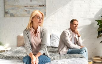 A woman and a man, both middle-aged, sit on a bed facing away from each other, looking pensive and distant. The room has a white brick wall, a painting, and some plants. Sunlight streams in, creating a stark, contemplative atmosphere.