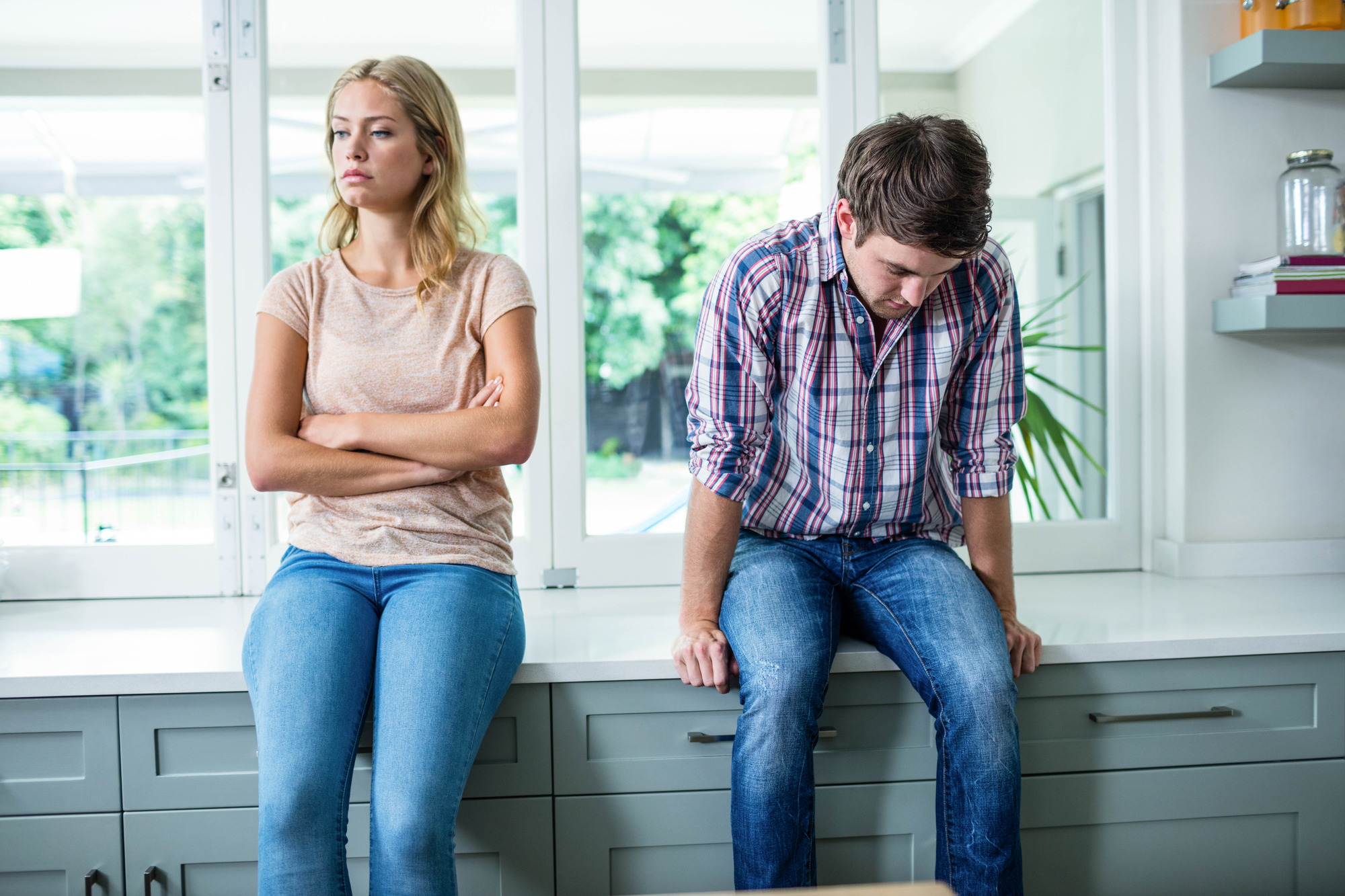 A woman and a man sitting on a kitchen counter, both facing forward. The woman has her arms crossed and looks away with a stern expression. The man has his hands on his lap, leaning forward slightly, looking down. The atmosphere appears tense.
