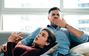 A man and woman are relaxing on a couch. The woman, in a maroon top, is lying with her head on the man's lap, pointing upwards seemingly explaining something. The man, in a blue shirt, is yawning with his hand covering his mouth. Light fills the room from large windows.