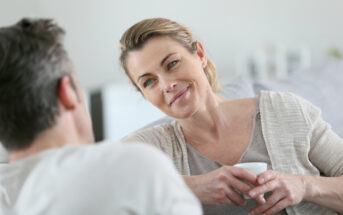 A woman with light brown hair tied back is sitting and smiling at a man who is facing away from the camera. She is holding a white mug with both hands and wearing a beige cardigan over a grey shirt. They appear to be in a relaxed, comfortable conversation.