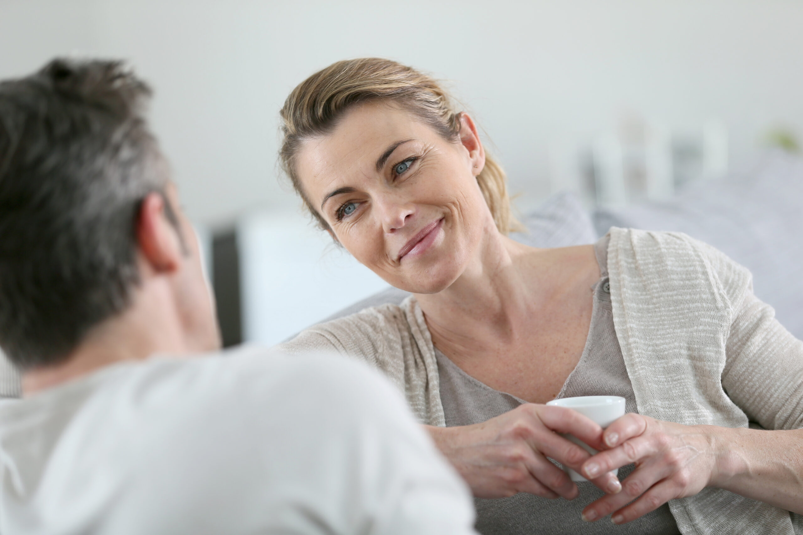 A woman with light brown hair tied back is sitting and smiling at a man who is facing away from the camera. She is holding a white mug with both hands and wearing a beige cardigan over a grey shirt. They appear to be in a relaxed, comfortable conversation.