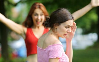 Two women are in an outdoor setting with trees in the background. One woman in a red top stands in the background with arms raised, while the woman in the foreground, wearing a pink off-shoulder top, appears upset or uncomfortable, with her hand near her face.