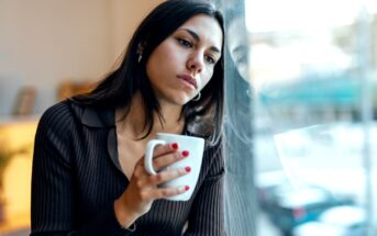 A woman with long dark hair sits by a window, holding a white mug. She looks contemplative and appears to be lost in thought, gazing out of the window. She is wearing a dark, long-sleeved top, and her fingernails are painted red.