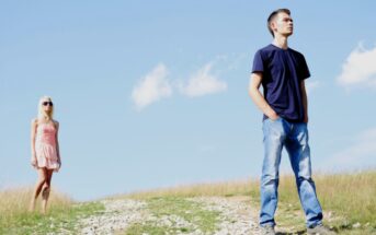 A man in a navy t-shirt and jeans stands with his hands in his pockets, looking to the right. A woman in a light pink dress and sunglasses stands in the background. They are in an open grassy field under a clear blue sky.