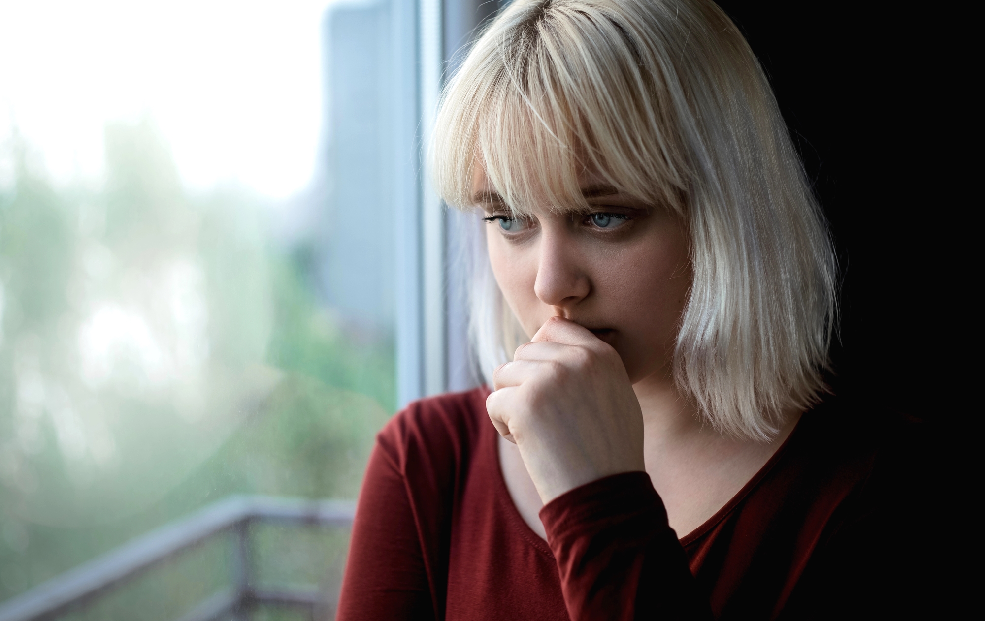 A young woman with blonde hair and bangs, wearing a maroon top, stands by a window with natural light filtering in. She gazes outside with a thoughtful expression, resting her chin on her hand. The background reveals a blurred outdoor view.