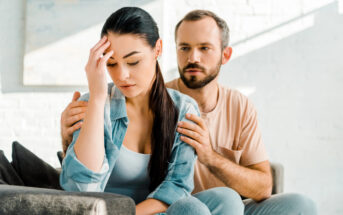 A woman sits on a couch looking distressed, with her hand on her forehead. A man sits beside her, gently placing his hands on her shoulders in a comforting manner. The scene appears to be well-lit with natural sunlight streaming in from a window.