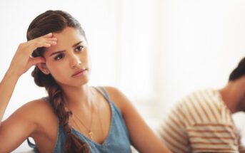 A woman with a long braid, wearing a blue sleeveless top, sits in the foreground with a thoughtful and concerned expression, resting her head on her hand. In the background, a person in a striped shirt is blurred and facing away. The background is light and minimalistic.