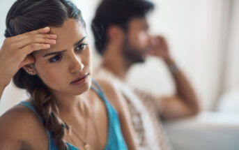 A woman with a concerned expression holds her forehead, sitting in the foreground. In the background, a man is blurred, appearing to be in distress or deep thought. Both are indoors, with soft lighting creating a somber mood.