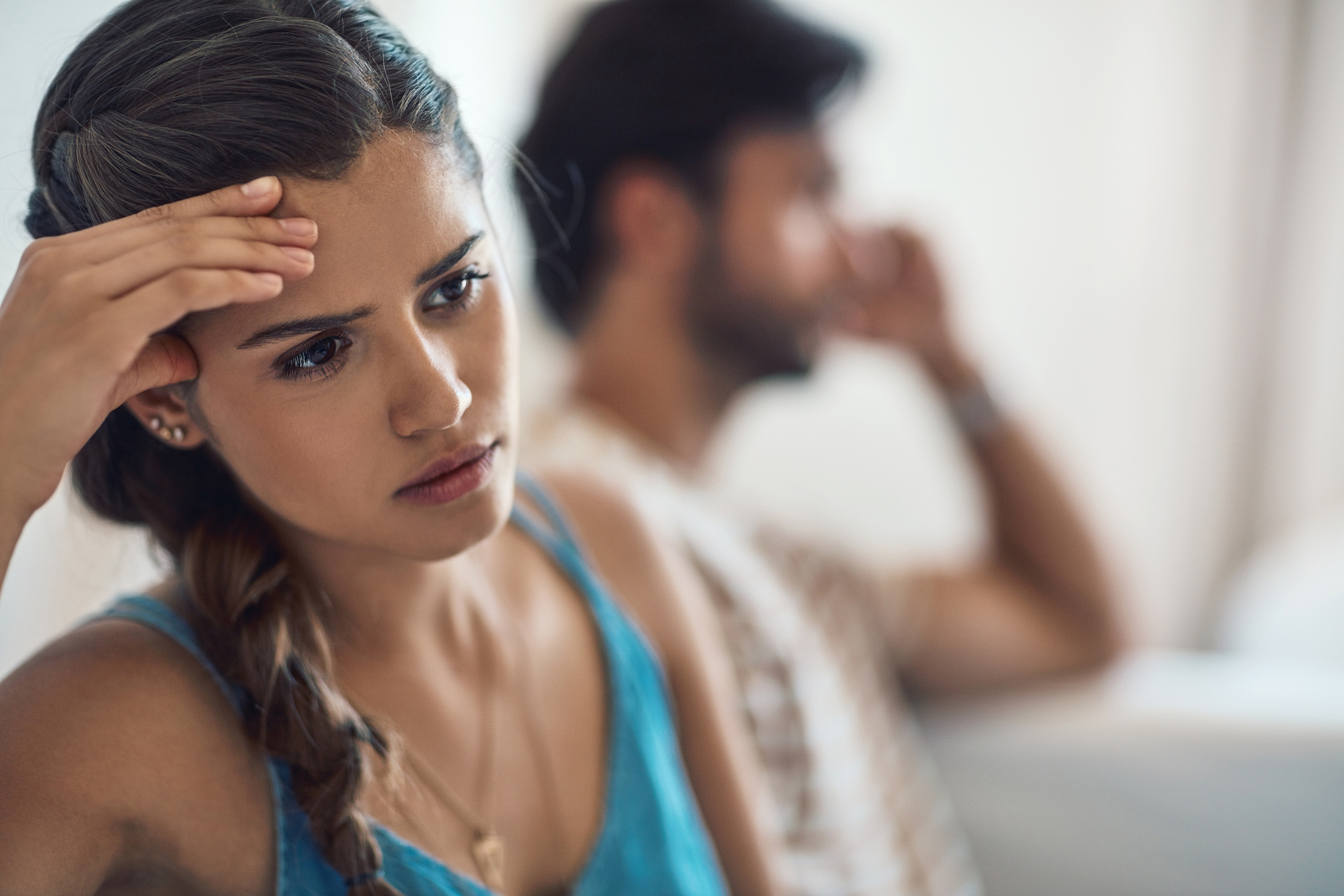 A woman with a concerned expression holds her forehead, sitting in the foreground. In the background, a man is blurred, appearing to be in distress or deep thought. Both are indoors, with soft lighting creating a somber mood.