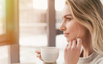 A blonde woman in a light sweater enjoys a warm drink from a white mug while gazing thoughtfully out of a window. Sunlight streams in, creating a serene and cozy atmosphere.