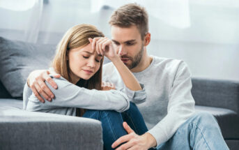 A man with a beard and short hair is sitting on a couch, comforting a woman with long brown hair who seems distressed. The woman has her eyes closed and is resting her forehead on her hand while the man has his arm around her shoulders. They both are wearing casual clothes.