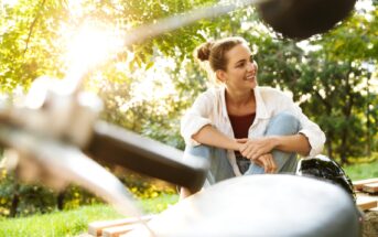 A woman with her hair in a bun sits outdoors under the trees, smiling and relaxed. She is wearing a white shirt over a dark top and light blue jeans. The sun is shining through the leaves, creating a warm, peaceful atmosphere. Nearby, there are blurred objects in the foreground.