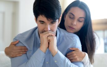 A person in a light blue shirt sits with a thoughtful expression and hands clasped near their face. Another person with long hair leans in from behind, gently embracing them and offering comfort. The scene suggests a moment of emotional support and empathy.