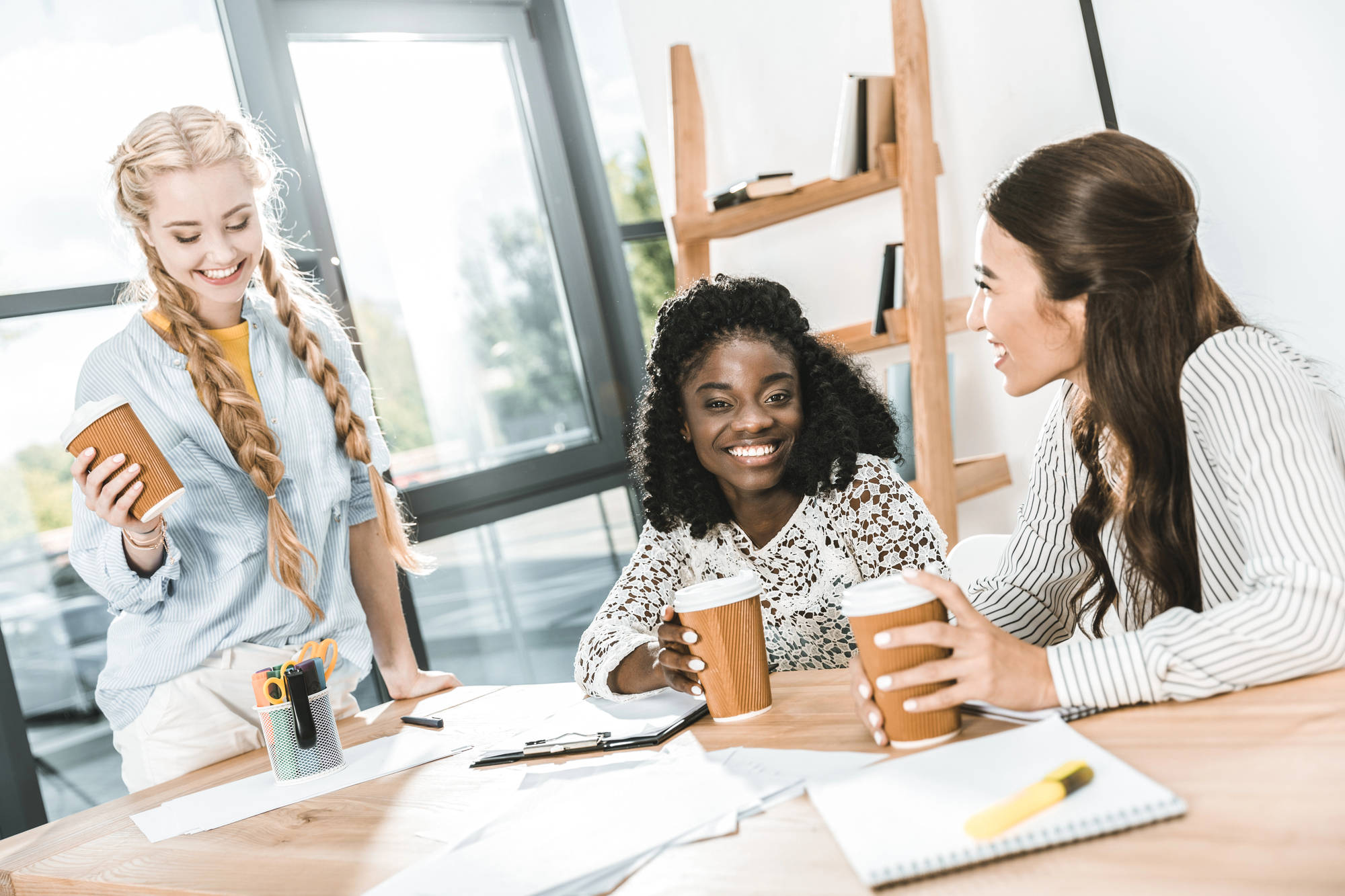 Three women are smiling and chatting around a table, holding coffee cups. They are in a bright room with large windows and wooden shelves in the background. Papers, pens, and a laptop are scattered on the table, indicating a casual work environment.