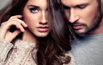 A close-up of a young woman and a man. The woman, on the left, has long brown hair, blue eyes, and is wearing a cozy beige sweater. The man, on the right, has short brown hair, a beard, and is wearing a gray sweater. Both have serious expressions.