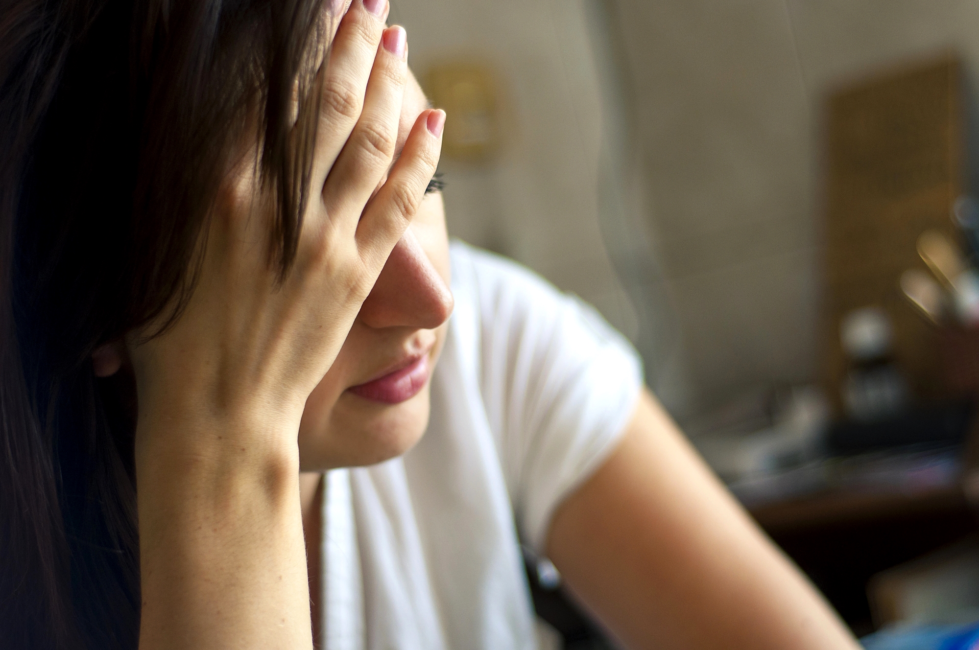 A woman with brown hair is shown resting her face in her hand, appearing distressed or tired. She is wearing a white shirt, and the background is blurred, indicating an indoor setting with indistinct objects.