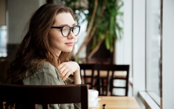 Woman with glasses and long, brown hair, sits at a wooden table in a cafe, looking out of a large window. She wears an olive green jacket and appears to be relaxed and thoughtful. In the background, there are chairs and a tall plant.