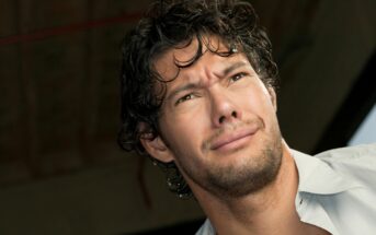 A man with curly dark hair and a light beard is looking off into the distance with a confused or concerned expression. He is wearing a light-colored shirt and is photographed from a low angle. The background is out of focus, suggesting an indoor setting.