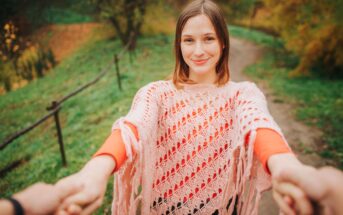 A smiling woman with light brown hair, wearing an orange top and a pink poncho, holds hands with someone while standing on a path in a park. The background features lush greenery and trees with autumn leaves.