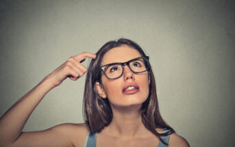 A woman with long brown hair and glasses is looking up and slightly to the side, appearing thoughtful. She is touching her head with her right hand, and her expression conveys that she is trying to remember something or is in deep thought. The background is plain and gray.