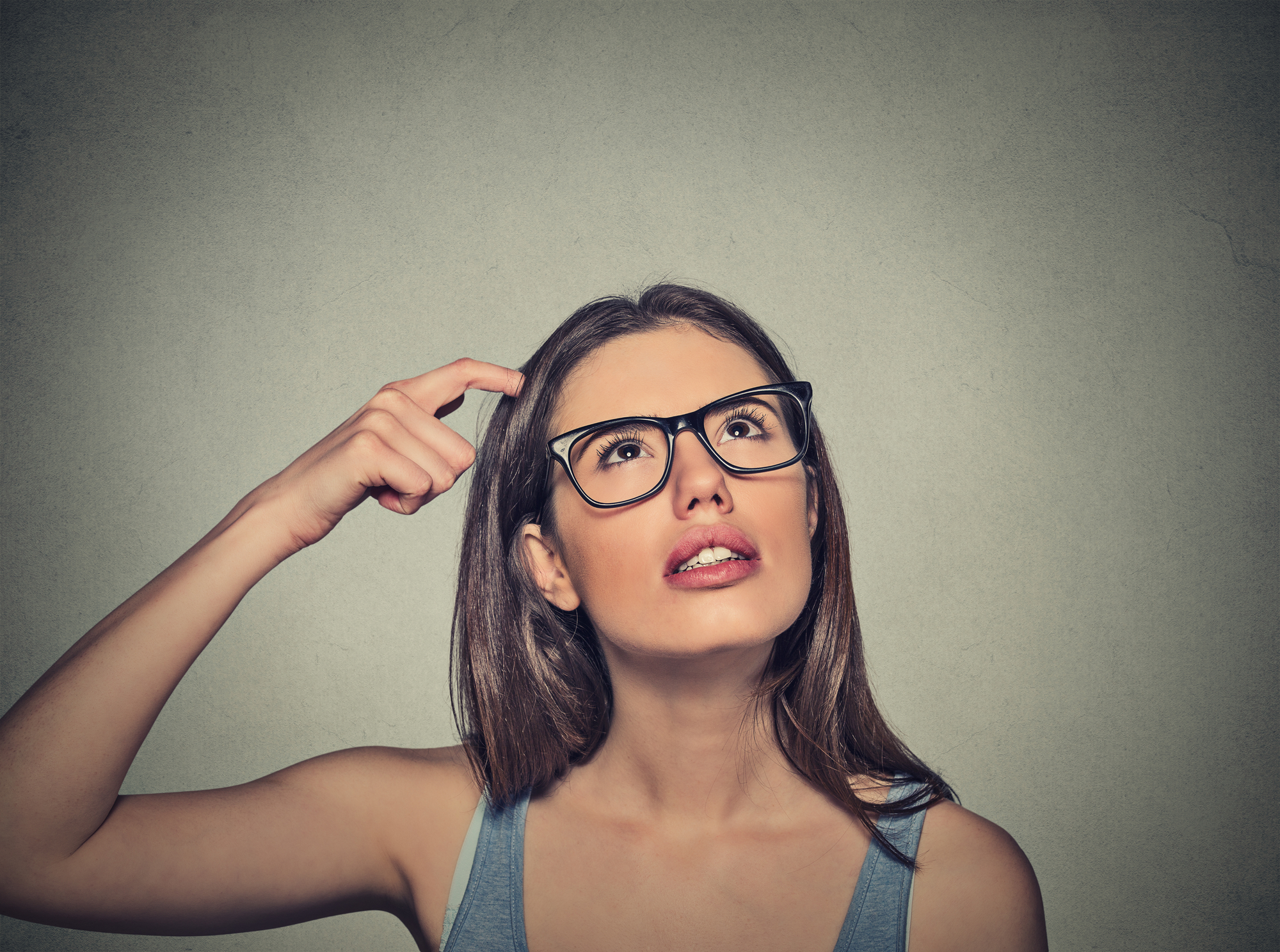 A woman with long brown hair and glasses is looking up and slightly to the side, appearing thoughtful. She is touching her head with her right hand, and her expression conveys that she is trying to remember something or is in deep thought. The background is plain and gray.